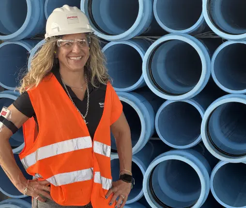 A woman in a hard hat, safety glasses, and orange reflective vest stands in front of stacked blue industrial pipes.